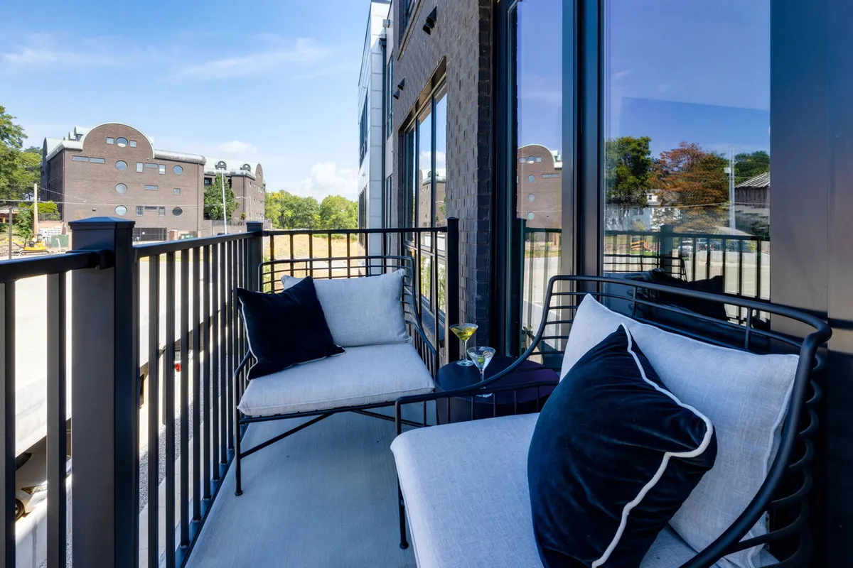 A private patio with two stylish chairs and a small table holding a drink, set against large windows. The tranquil atmosphere suggests a perfect spot for morning coffee or evening relaxation, with hints of urban sophistication and privacy.