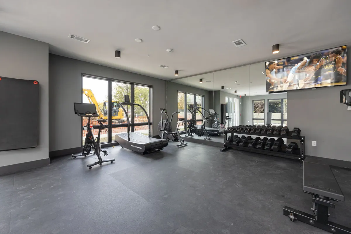 A modern, well-lit gym featuring a treadmill, weight rack, and large windows. The space feels active and motivating, ideal for solo workouts, personal training, or group fitness classes. A television screen adds entertainment for residents while they exercise.