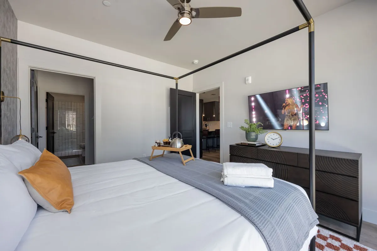 A canopy king bed takes center stage in this inviting bedroom, flanked by a nightstand with a tray and a smart TV mounted on the wall. The room features warm, cozy lighting ideal for relaxation and entertainment, highlighting the suite's plush bedding and tranquil atmosphere.