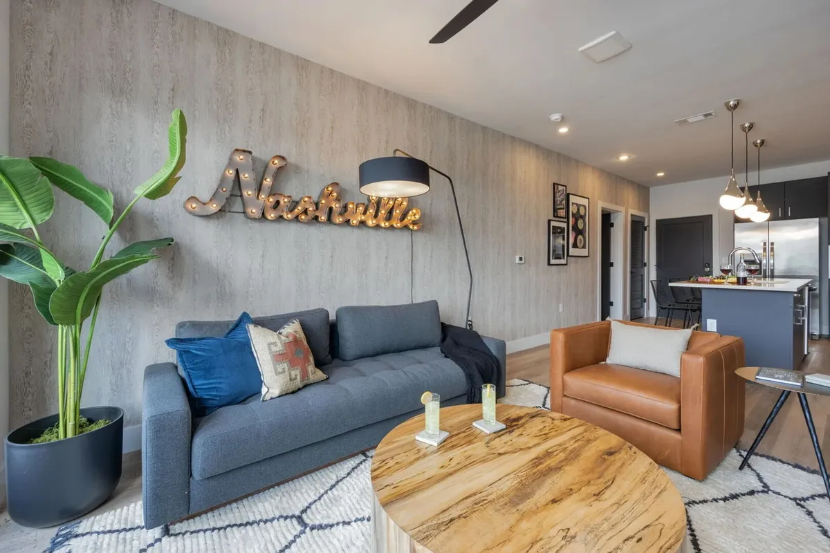 A cozy living room featuring a modern sofa, coffee table, armchair, and a decorative 'Nashville' sign on the wall, accented by lush green plants. The arrangement is inviting, with ample space for socializing or relaxing, and the decor ties into the local character.