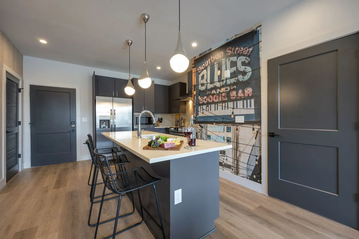 This image features a sophisticated kitchen interior with a spacious island countertop, modern appliances, hanging light fixtures, and a vintage mural accent. The island includes bar seating, and ample space is available for both meal prep and entertaining guests.