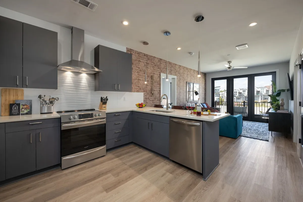 A modern kitchen interior showcasing a large kitchen island, stainless steel appliances, an exposed brick wall, and large windows offering an outdoor view. The expansive island is ideal for prepping meals and seating multiple guests.