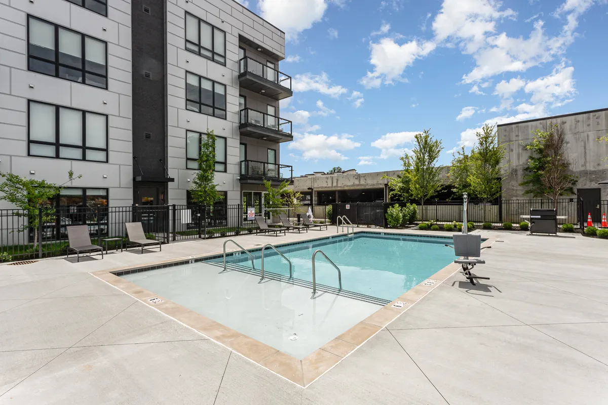 A sunny, relaxed exterior view of a resort-style swimming pool flanked by lounge chairs and surrounded by green landscaping and the modern condo building. The inviting pool area is ready for swimming, sunbathing, or social gatherings, with a family-friendly vibe.