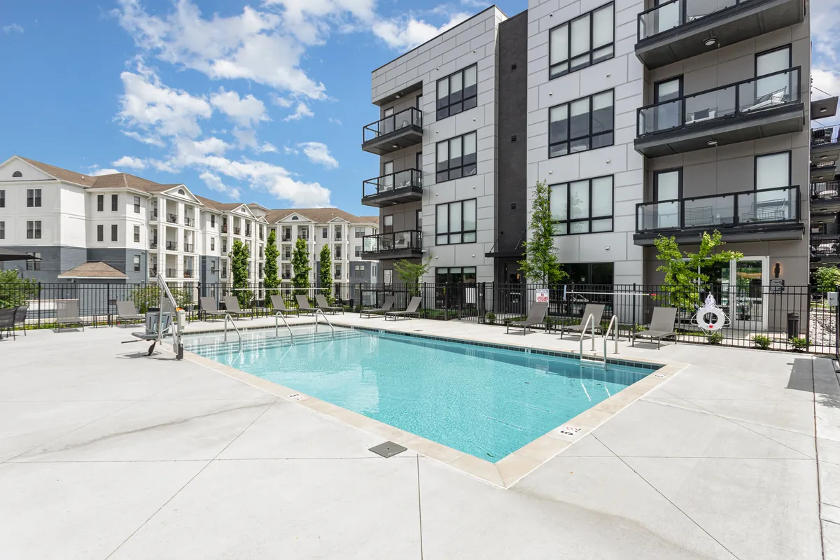A sparkling swimming pool surrounded by landscaped greenery, with balcony views above. The scene radiates relaxation with people swimming, sunbathing, and gathering for social events, evoking the feel of a high-end resort.